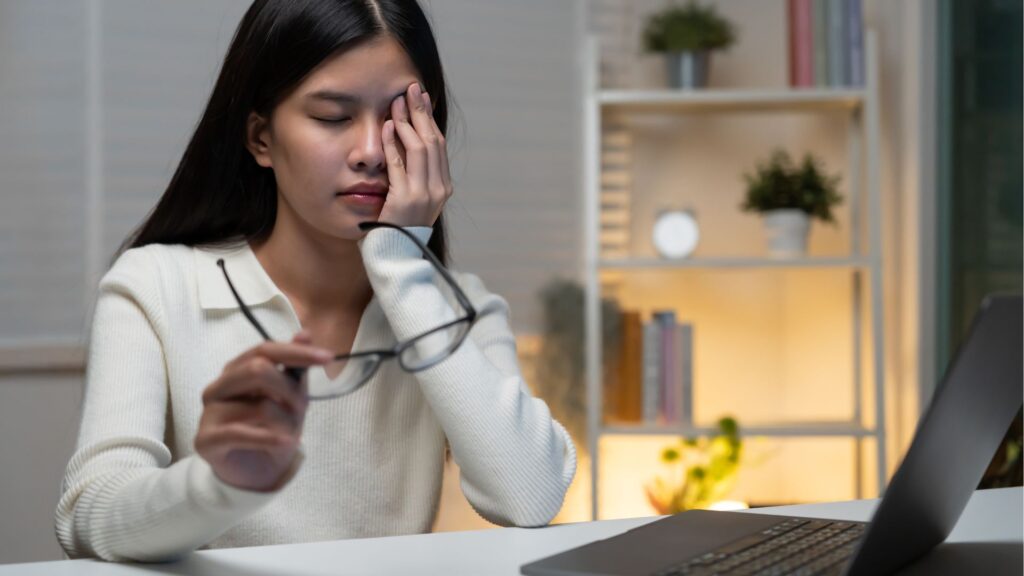 デスクワーク中に目の疲れやかすみを感じ、辛そうに目頭を押さえる女性。糖分の摂りすぎが引き起こす眼精疲労や視界のぼやけのイメージ。