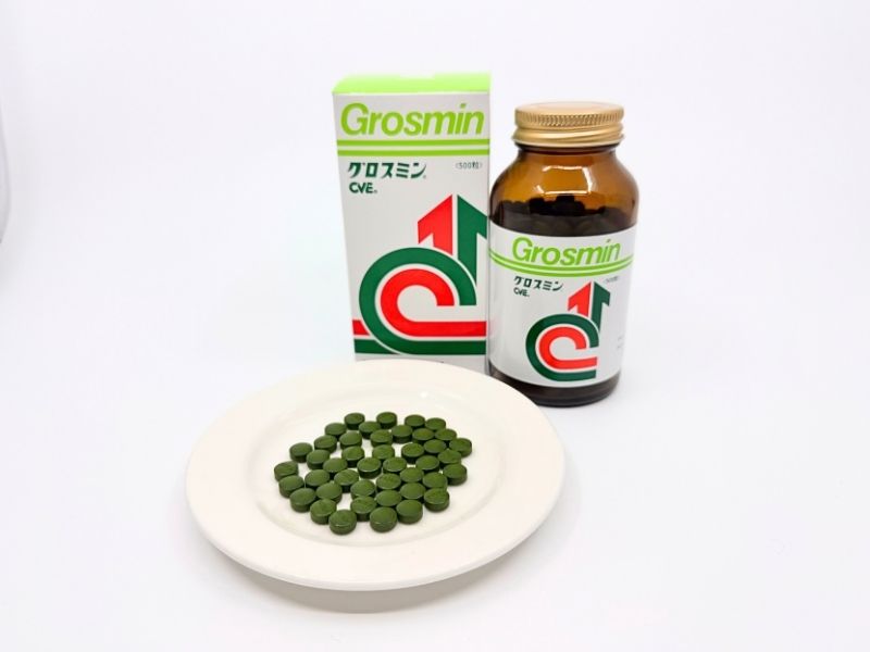 グロスミンのパッケージと瓶と粒。緑と白の箱にCVEのロゴ、琥珀色のガラス瓶。白い皿に並べた緑色の丸い粒。