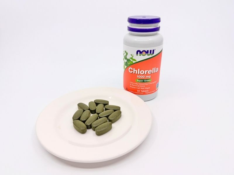 NOW Foods Chlorella 1000mgのボトルと粒。白いボトルにオレンジのラベル。白い皿に並べたカプセル型の大きな緑色の粒。