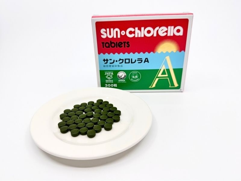 サン・クロレラAのパッケージと粒。赤い箱にJHFA認定マークとインフォームドチョイスのロゴ。白い皿に並べた深緑色の丸い粒。