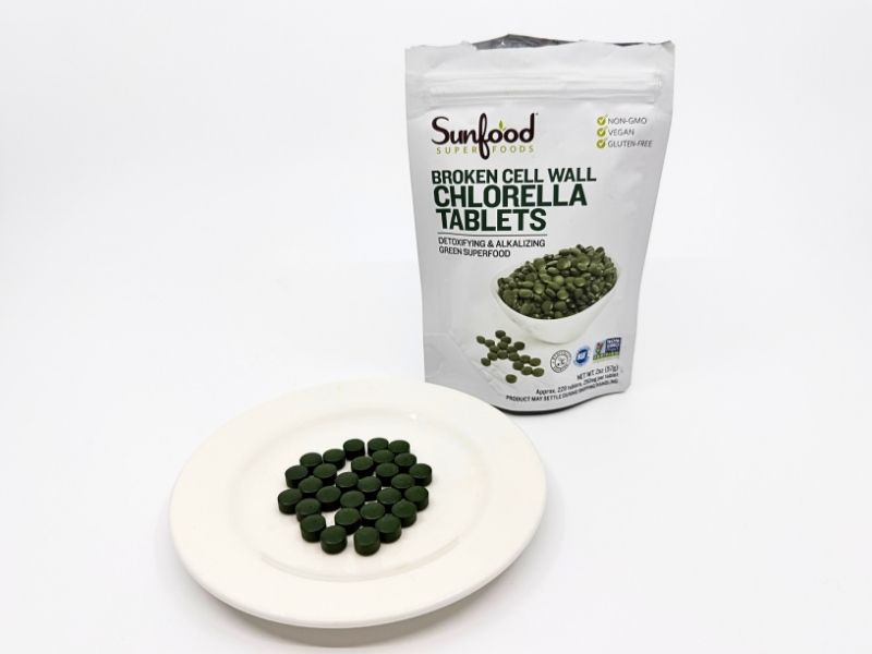 unfood Broken Cell Wall Chlorella Tabletsのパウチと粒。白いパウチにNON-GMO・VEGAN・GLUTEN FREEの表示。白い皿に並べた小さな深緑色の丸い粒。