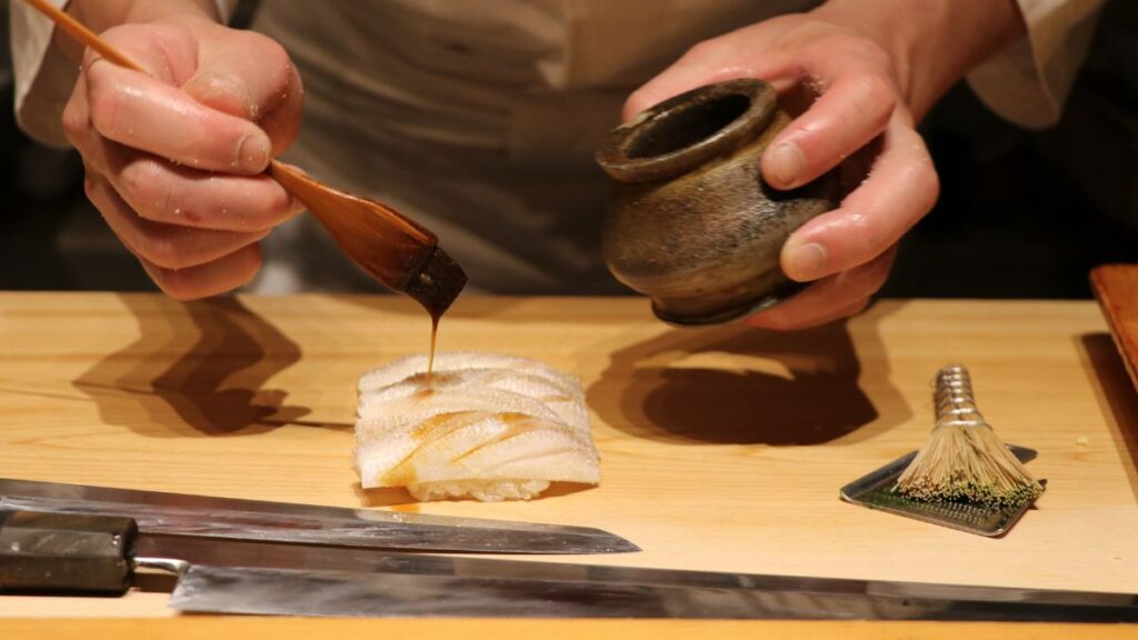 「江戸前鮨の職人が、煮切り醤油をハケでネタに塗っている様子。本物の鮨屋では、客が自分で醤油をつけることはない。」